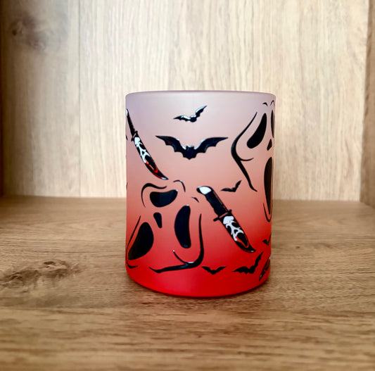 A frosted glass mug with a Halloween design, featuring a red-to-white gradient and black bat and dagger motifs, set on a wooden surface against a wooden background.