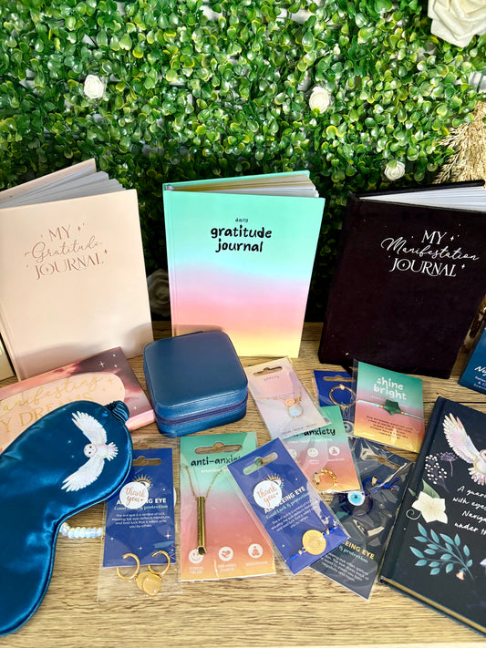 A styled flat-lay display of various self-care and journaling products, including multiple journals, a blue satin sleep mask, a small blue jewelry box, and several packaged earrings and necklaces, arranged on a wooden surface in front of a green foliage wall.