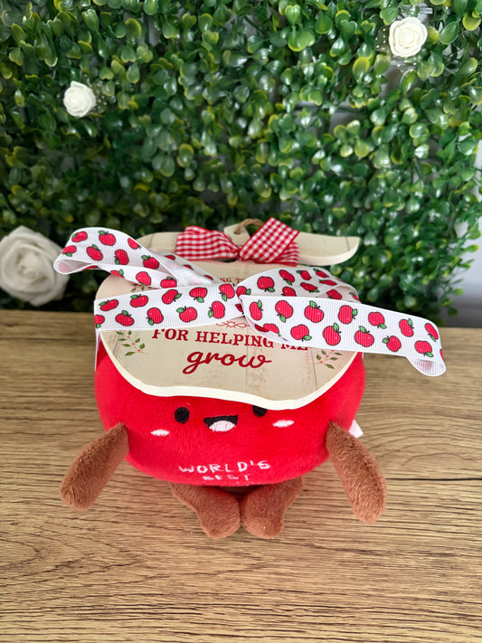 apple plush toy with brown arms and legs, with wooden apple sign tied on top with apple-printed ribbon on a wooden surface with green leafy background.