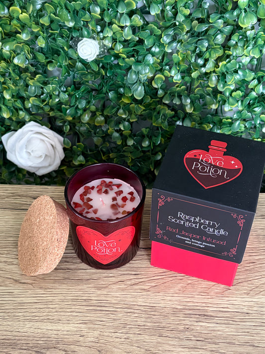 A Love Potion Raspberry Scented Candle shown with its black and red packaging box, alongside a cork lid, set against a green leafy background on a wooden surface.