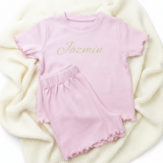 A girl's two-piece lounge set in light pink, featuring a short-sleeve top with 'Jazmin' written in gold cursive and matching ruffled shorts.
