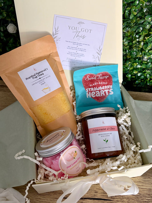 A curated gift box containing bath potion in kraft bag, sweets, whipped soap and aromatherapy candle jar on a wooden surface in ivory coloured gift box.