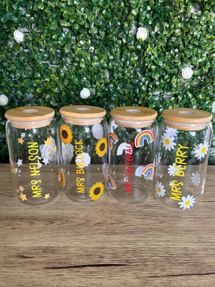 Four clear glass jars with light wood lids, decorated with personalised names and colourful sunflower, daisy, rainbow, and unicorn prints, arranged on a wooden surface in front of a green leafy wall.
