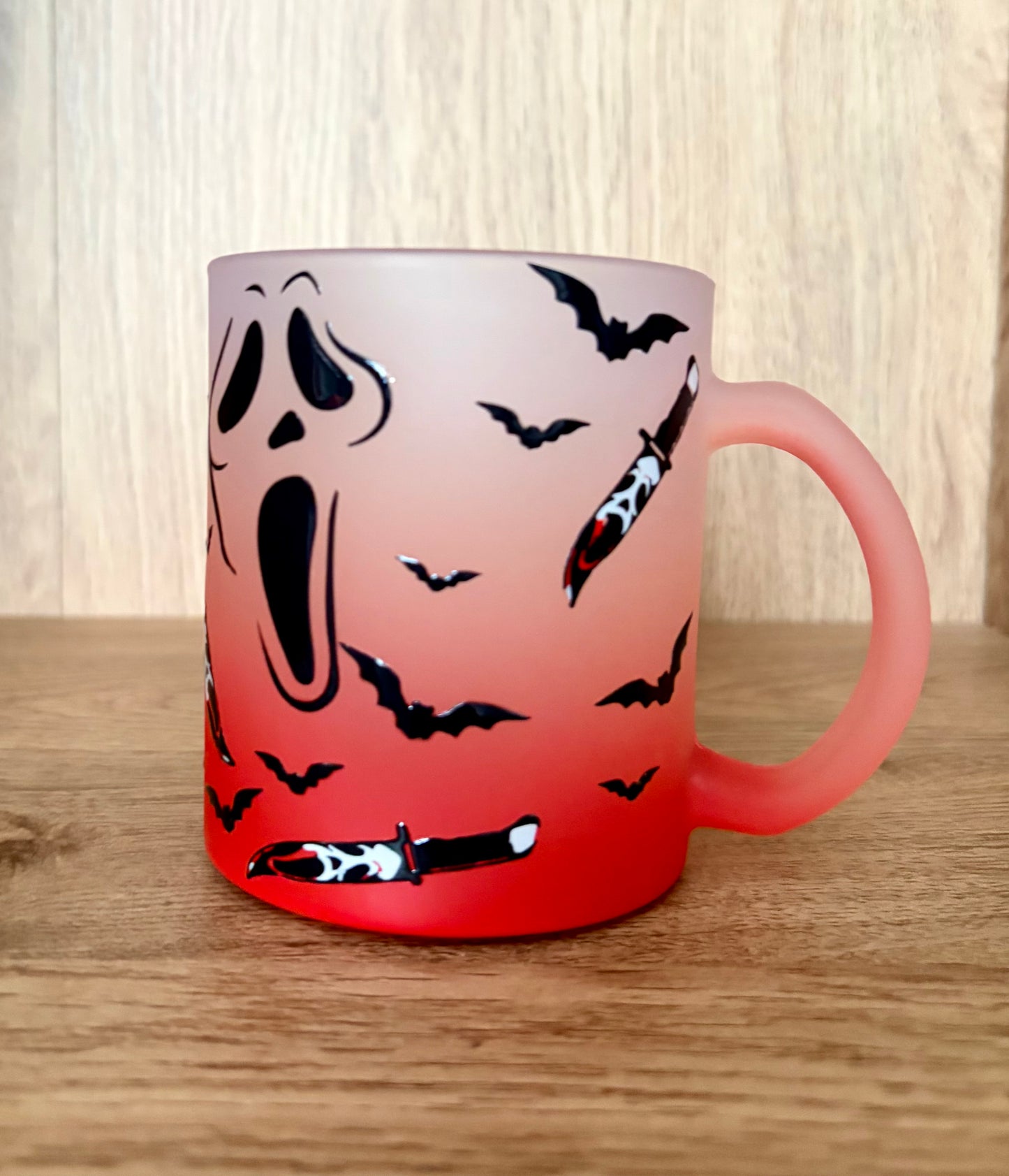 A frosted glass mug with a Halloween design, featuring a Ghostface face, black bats, and cutout knives over a red-to-white gradient, set on a wooden surface.