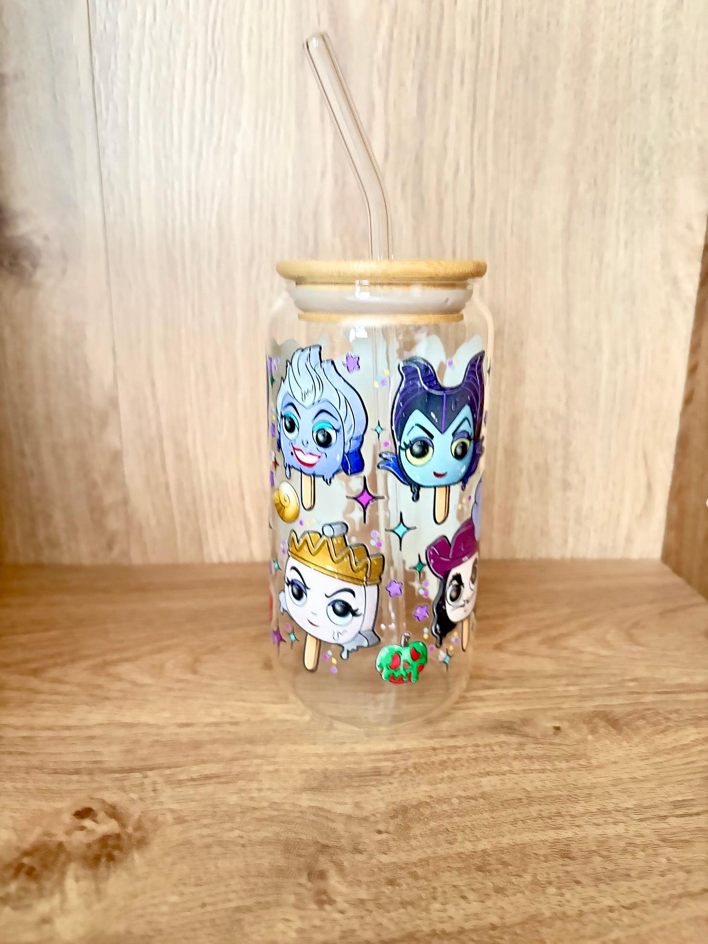 A clear glass can-shaped tumbler with a light wood lid and clear straw, decorated with cartoon ice cream characters on a wooden surface against a wood-paneled background.