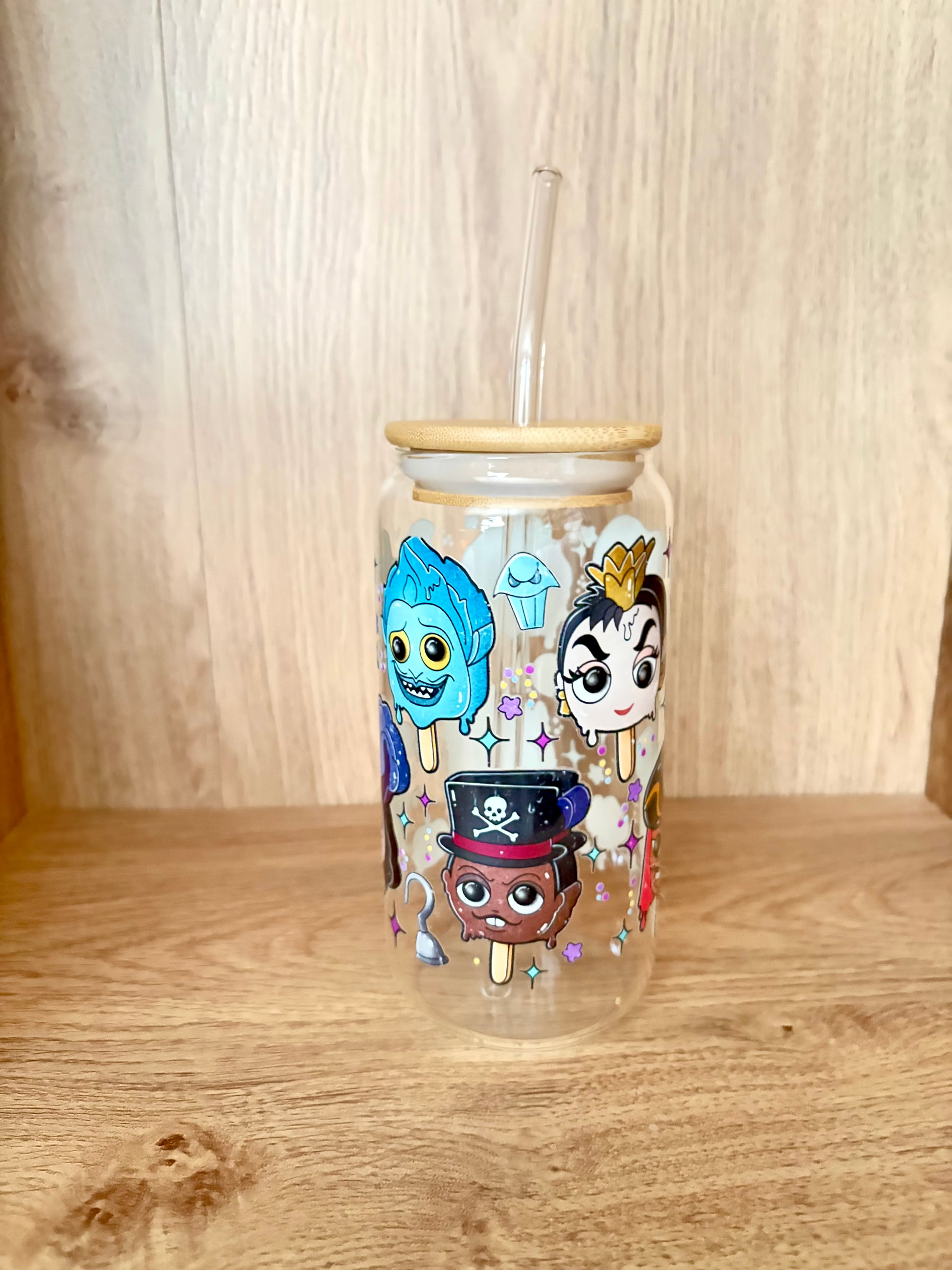 A clear glass tumbler with a bamboo lid and clear straw, decorated with colorful cartoon characters and ice cream cone illustrations, set on a wooden surface against a wooden background.