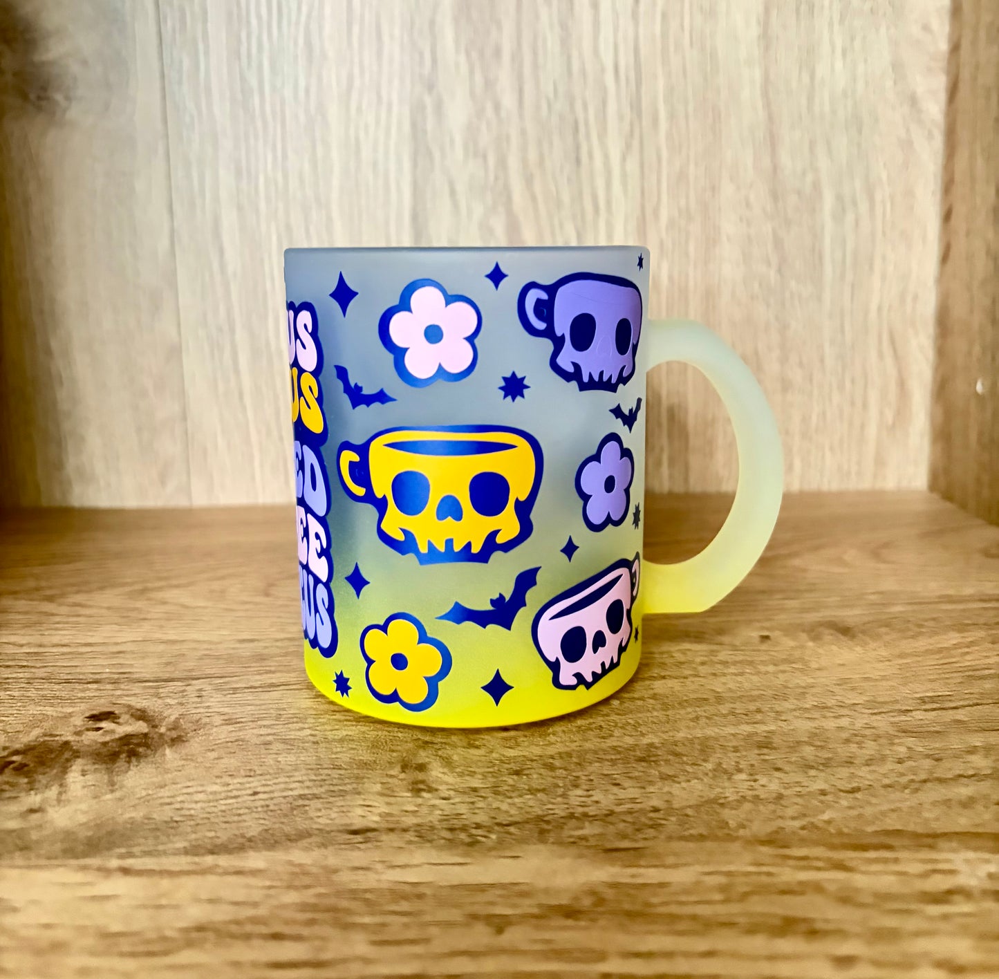 A frosted glass mug with a colorful Halloween-themed print of skulls, teacups, bats, flowers,' in yellow and blue, sitting on a wooden surface.
