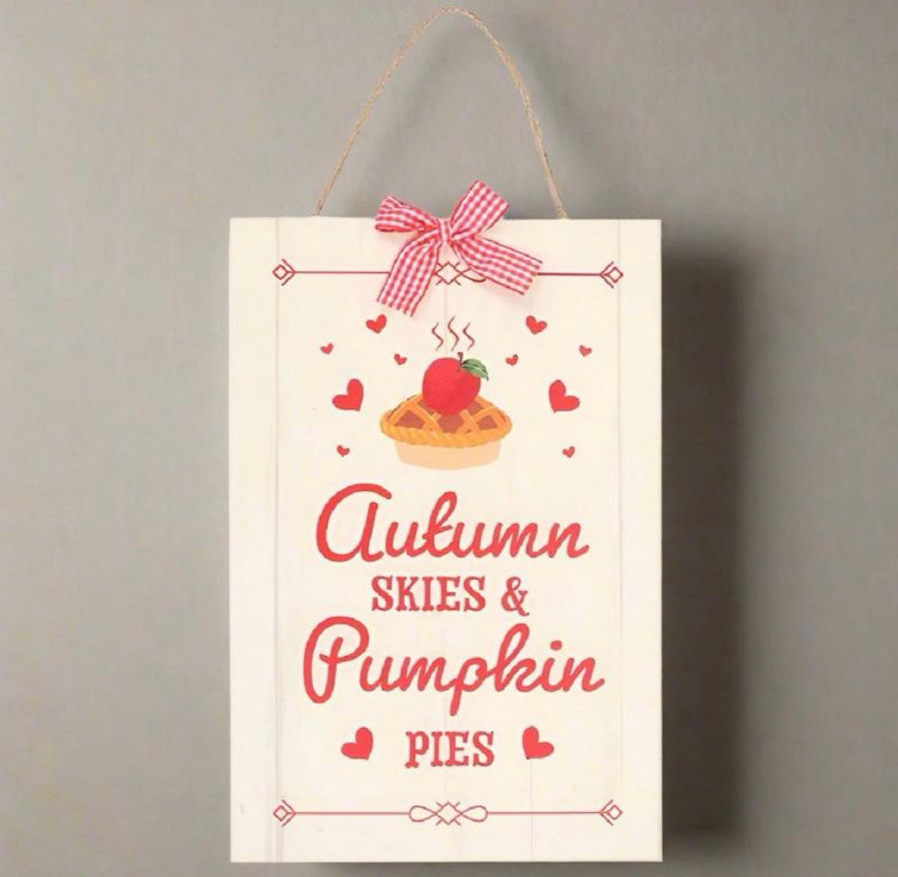 A vertical white wooden hanging sign with a twine loop at the top, decorated with a red-and-white gingham bow, a pumpkin pie illustration, and the words 'Autumn Skies & Pumpkin Pies' in red script and serif lettering, shown against a plain gray wall.