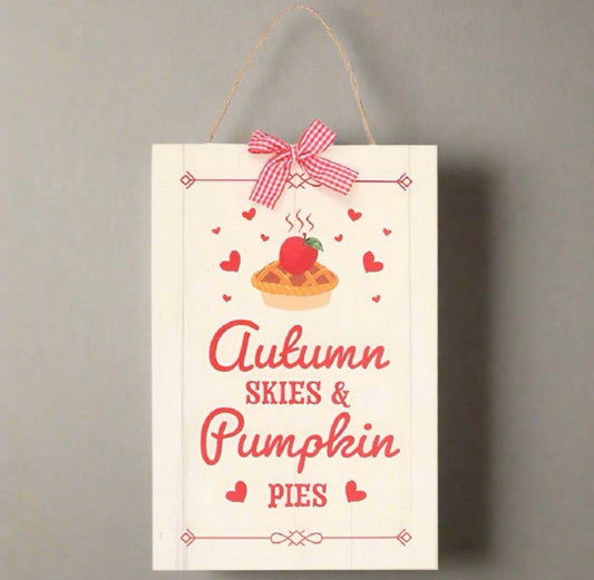 A vertical white wooden hanging sign with a twine loop at the top, decorated with a red-and-white gingham bow, a pumpkin pie illustration, and the words 'Autumn Skies & Pumpkin Pies' in red script and serif lettering, shown against a plain gray wall.