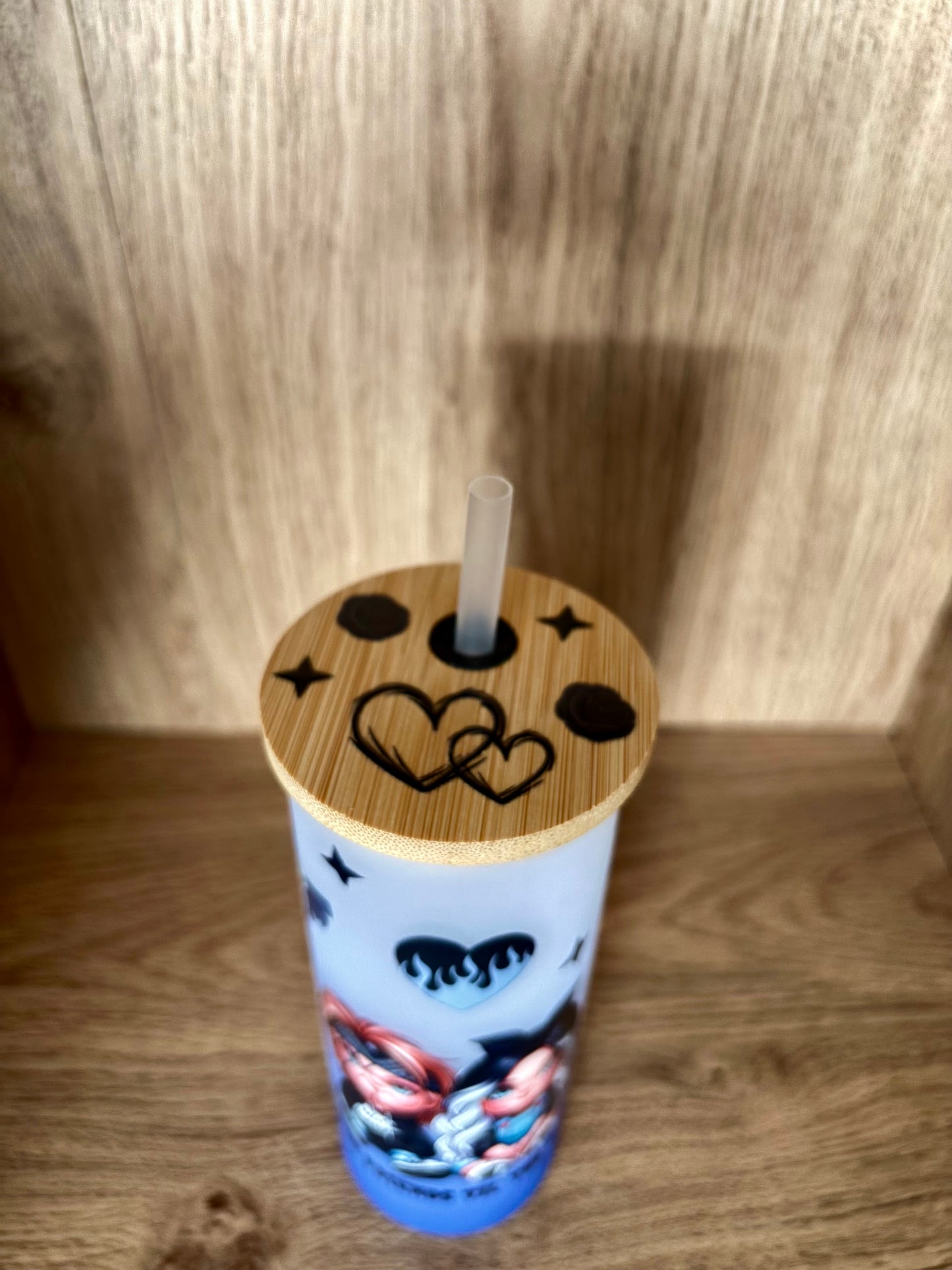 A light blue insulated tumbler with a bamboo lid and clear straw, decorated with black moon and heart graphics and a colorful character illustration near the bottom, set against a wooden background.