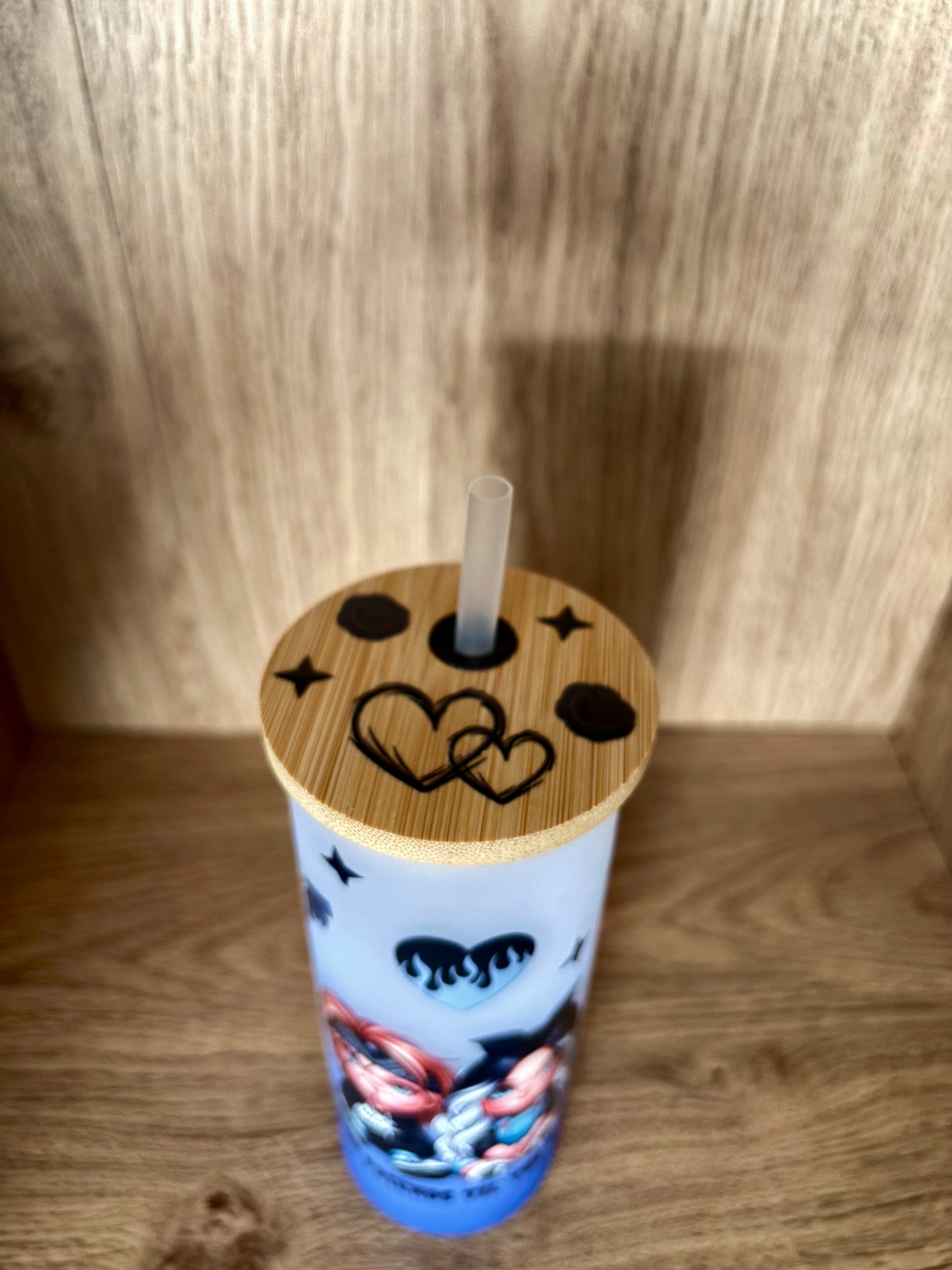 A light blue insulated tumbler with a bamboo lid and clear straw, decorated with black moon and heart graphics and a colorful character illustration near the bottom, set against a wooden background.