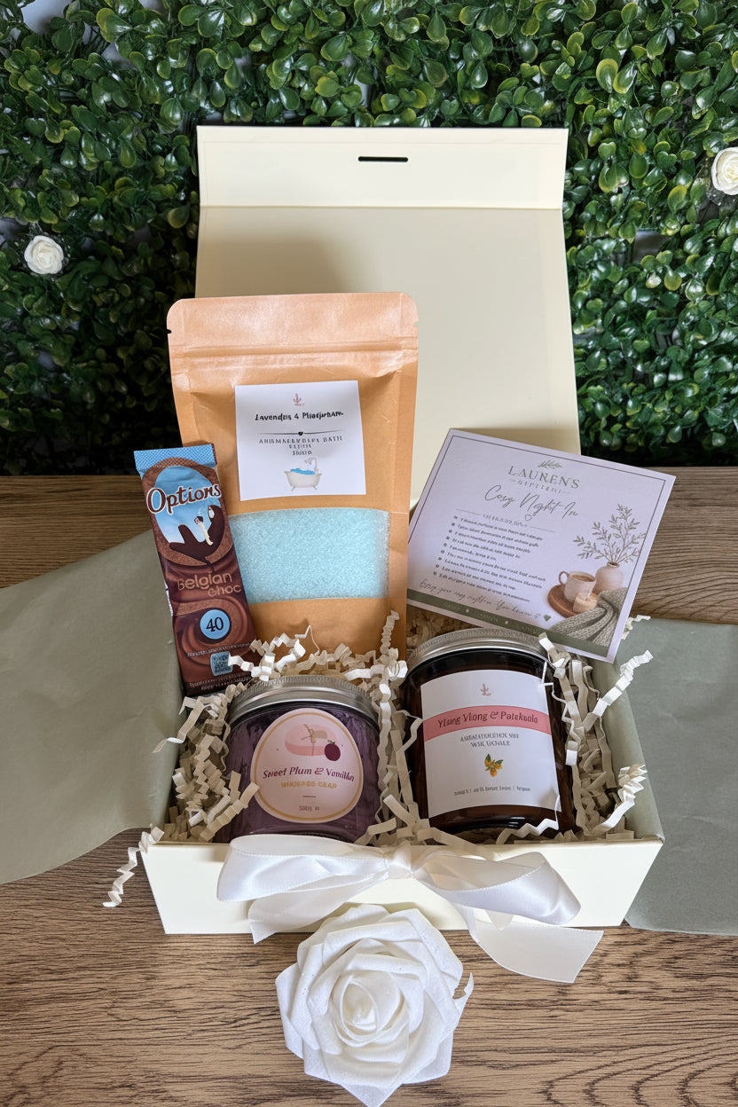 curated gift set containing bath salts, whipped soap, aromatherapy candle, hot chocolate and cosy night in affirmations card in an ivory gift box on a wooden surface with a green leafy backdrop
