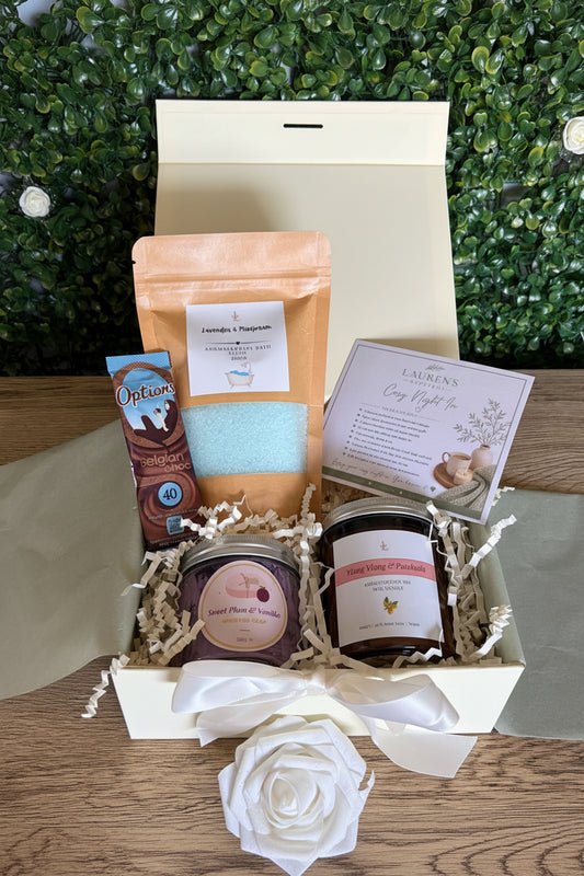 curated gift set containing bath salts, whipped soap, aromatherapy candle, hot chocolate and cosy night in affirmations card in an ivory gift box on a wooden surface with a green leafy backdrop