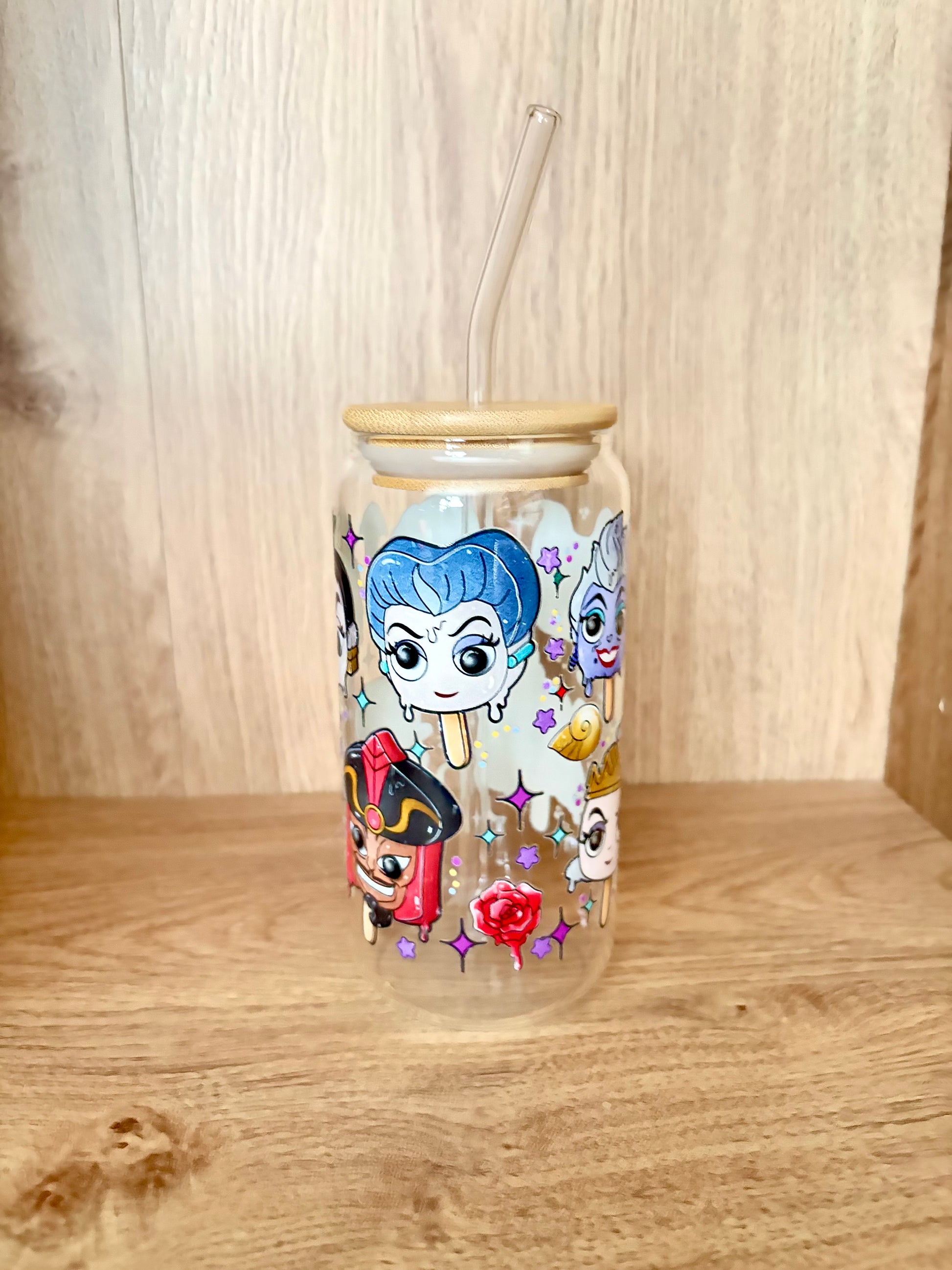 A clear glass tumbler with a bamboo lid and clear glass straw, decorated with colorful cartoon characters and sparkles, set on a wooden surface against a wooden background.