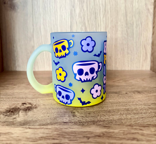 A frosted glass mug with a pastel blue-to-yellow gradient, decorated with blue skull teacups, yellow flowers, and blue bats, sitting on a wooden surface.
