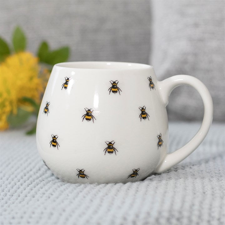Round white ceramic mug with all-over illustrated bumblebee print, styled with yellow flowers in the background