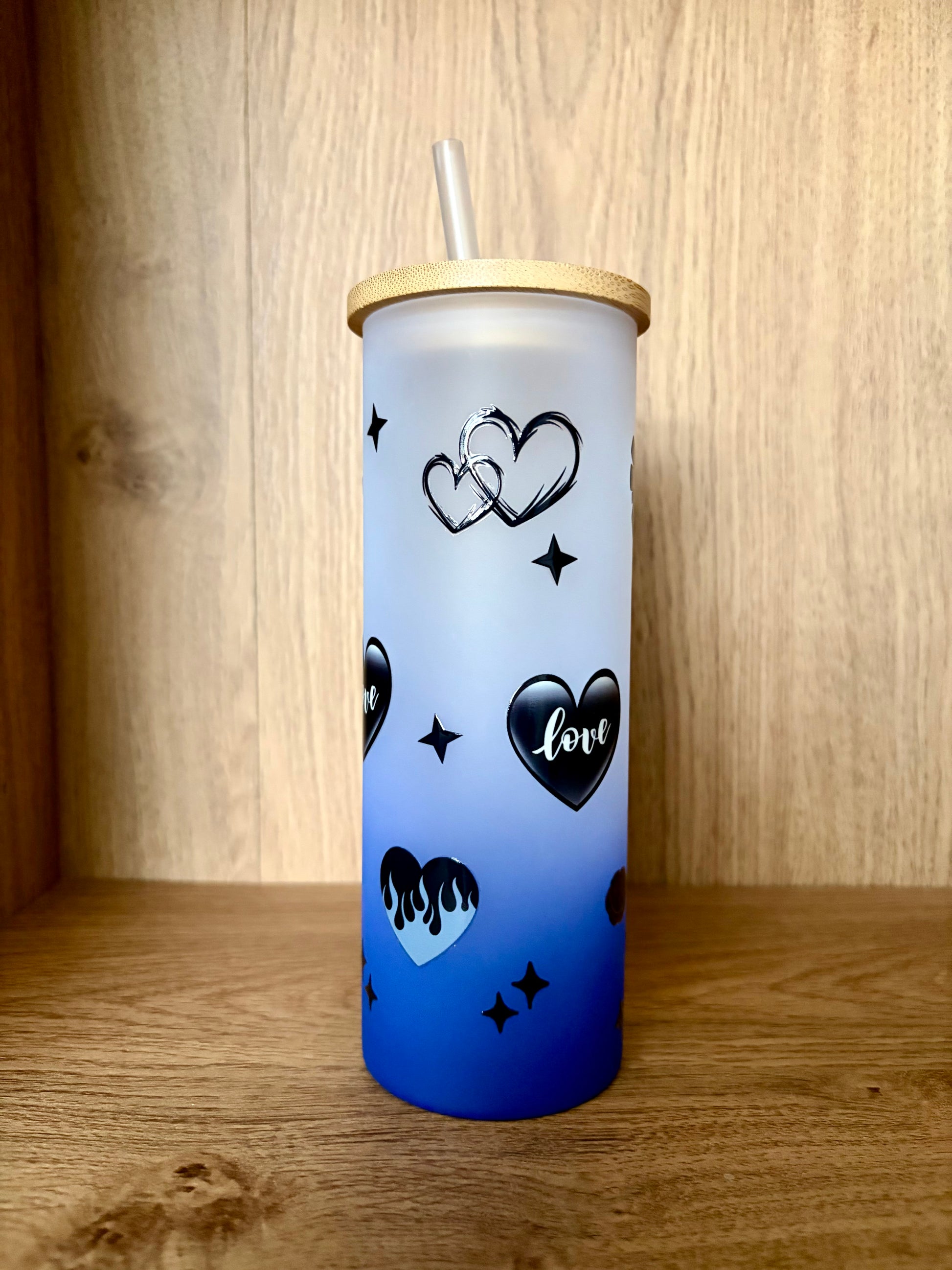 A frosted tumbler with a blue gradient base, decorated with black heart designs, stars, and the word 'love', featuring a bamboo lid and clear straw, set against a wooden background.