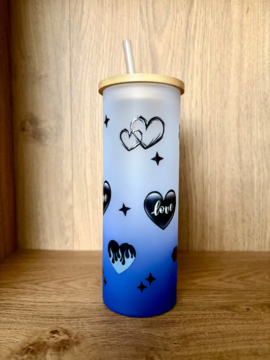 A frosted tumbler with a blue gradient base, decorated with black heart designs, stars, and the word 'love', featuring a bamboo lid and clear straw, set against a wooden background.