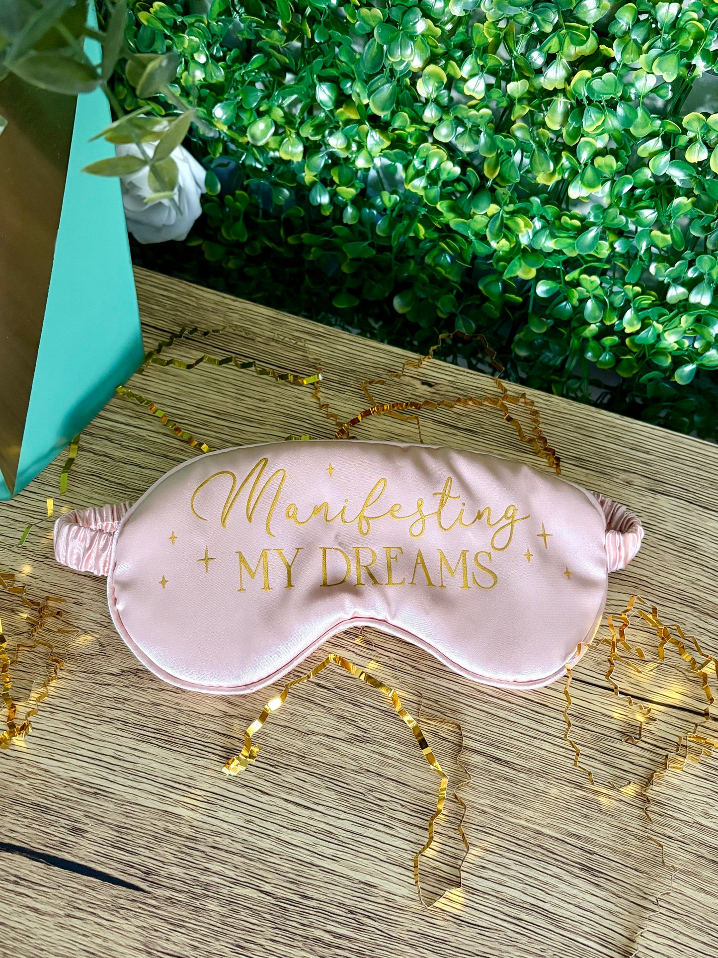 A light pink satin sleep mask with gold foil text reading 'Manifesting MY DREAMS' and small gold stars, laid on a wooden surface with decorative gold tinsel and green foliage in the background.