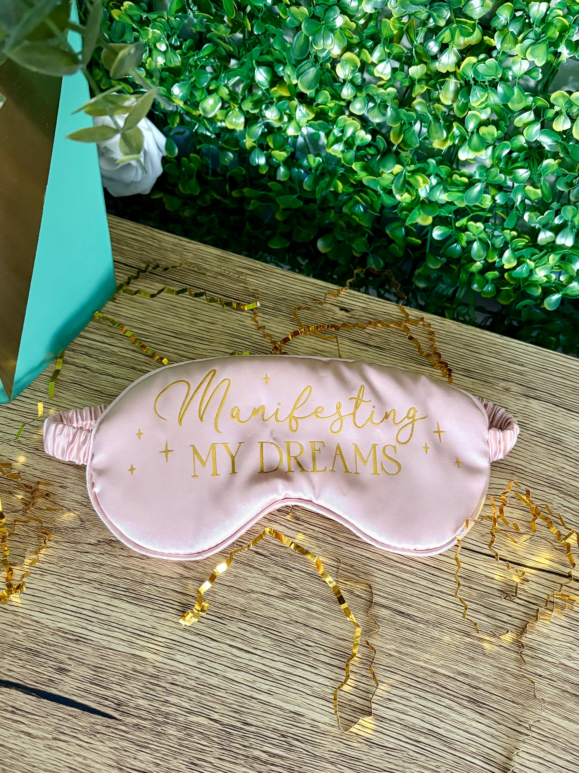 A light pink satin sleep mask with gold foil text reading 'Manifesting MY DREAMS' and small gold stars, laid on a wooden surface with decorative gold tinsel and green foliage in the background.