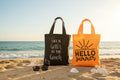Black tote bag with 'Life is better at the beach' text and orange tote bag with 'Hello summer' text on a grid background