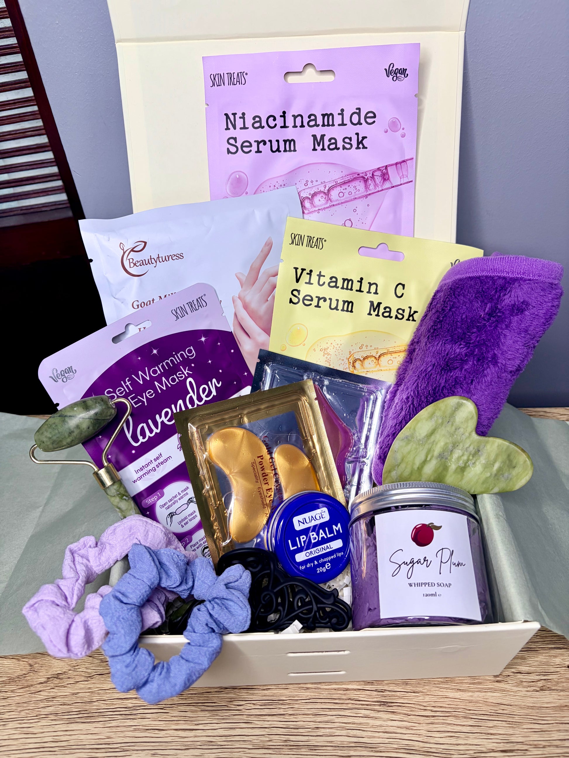 A curated skincare gift set arranged in a white tray, featuring multiple branded product packages including Niacinamide Serum Mask, Vitamin C Serum Mask, Lavender Eye Mask, Sugar Plum whipped soap, and a purple face cloth, alongside scrunchies and a green jade roller on a light wood surface.