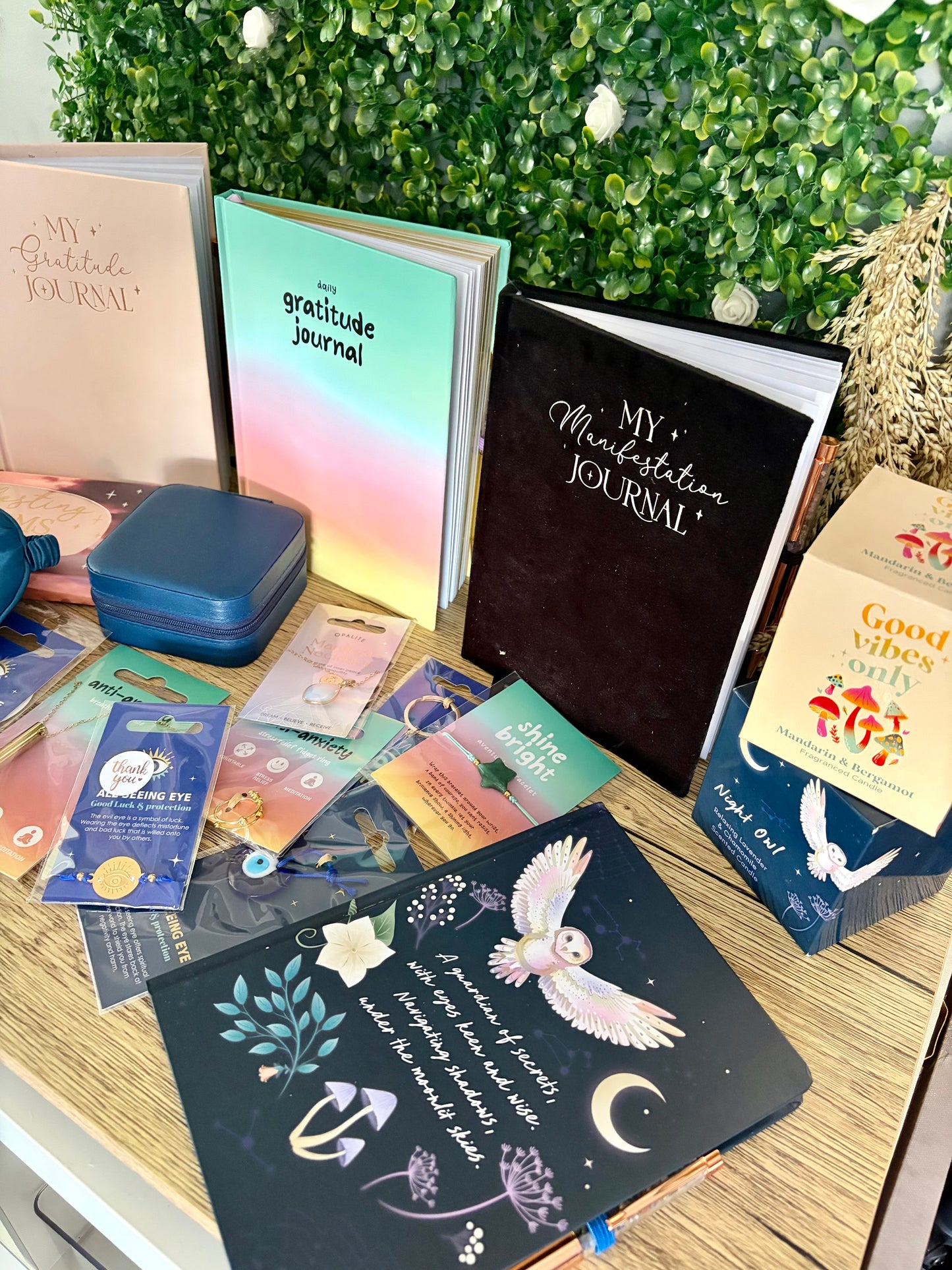 A styled tabletop display of multiple journaling and reading products, including notebooks, a gratitude journal, a black 'My Manifestation Journal,' cards, a blue case, and a 'Good Vibes Only' candle, arranged in front of a green foliage backdrop.