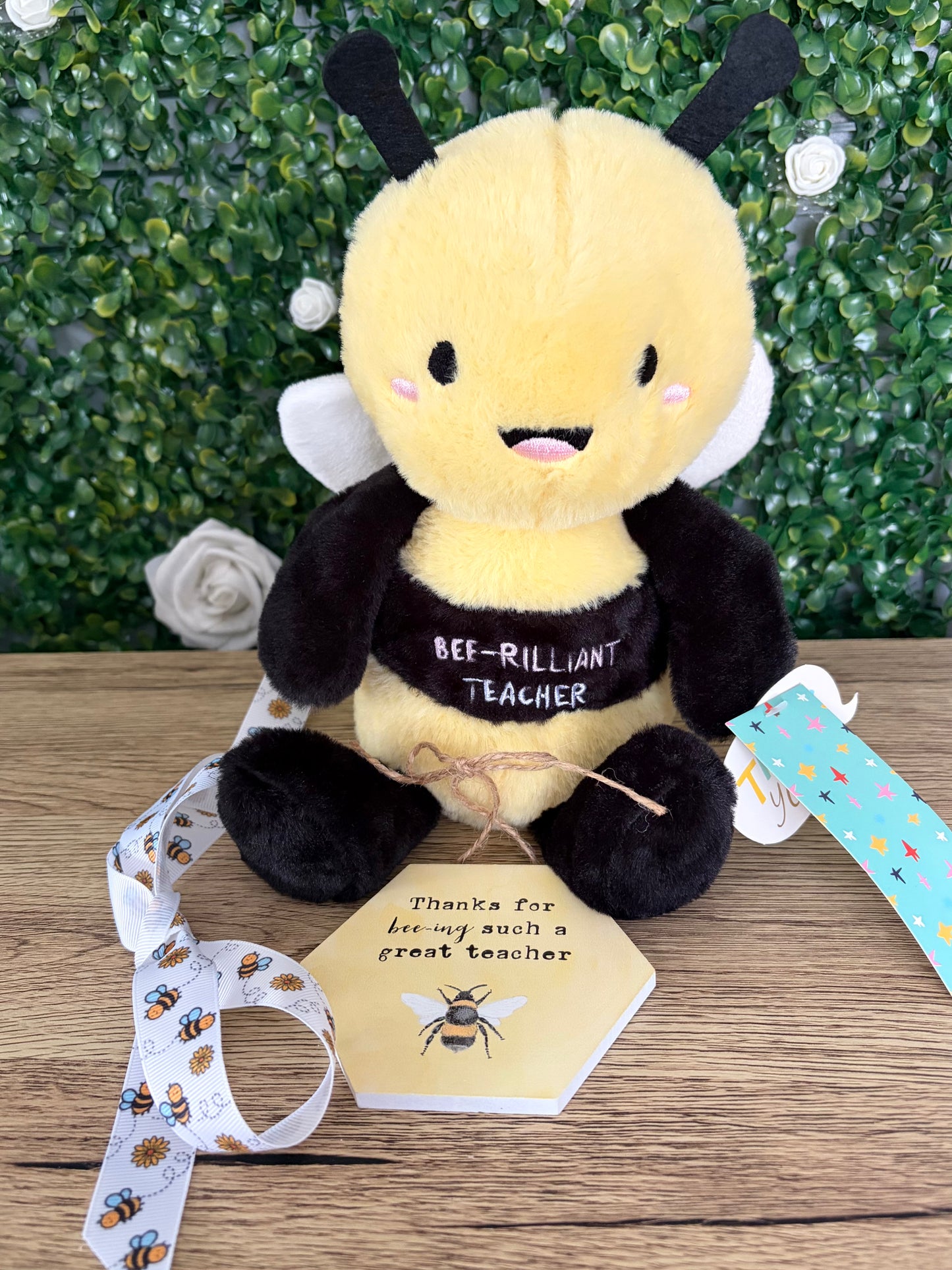 A plush bee toy sits on a wooden surface with a green hedge backdrop. The toy is yellow and black with white wings and the words 'BEE-ILLIANT TEACHER' on its belly.