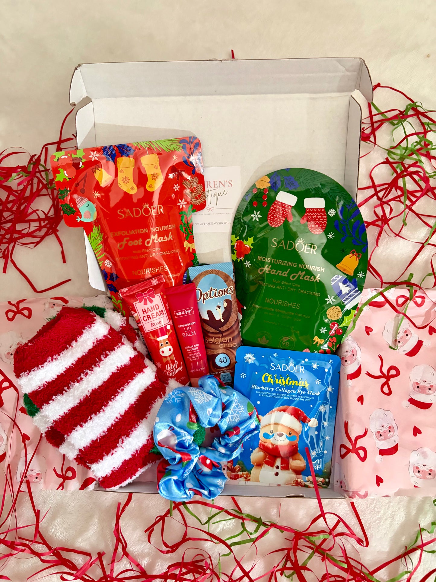 A curated festive self-care box filled with a variety of items such as cosy socks, hand cream, lip balm, eye patches, hand mask, foot mask, hair scrunchie & hot chocolate sachet
