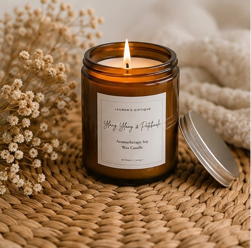 An amber glass jar candle with a lit flame sits on a woven straw mat, with dried flowers on the left and a silver lid resting beside it. The front label reads Lauren's Giftique and Ylang Ylang & Patchouli.