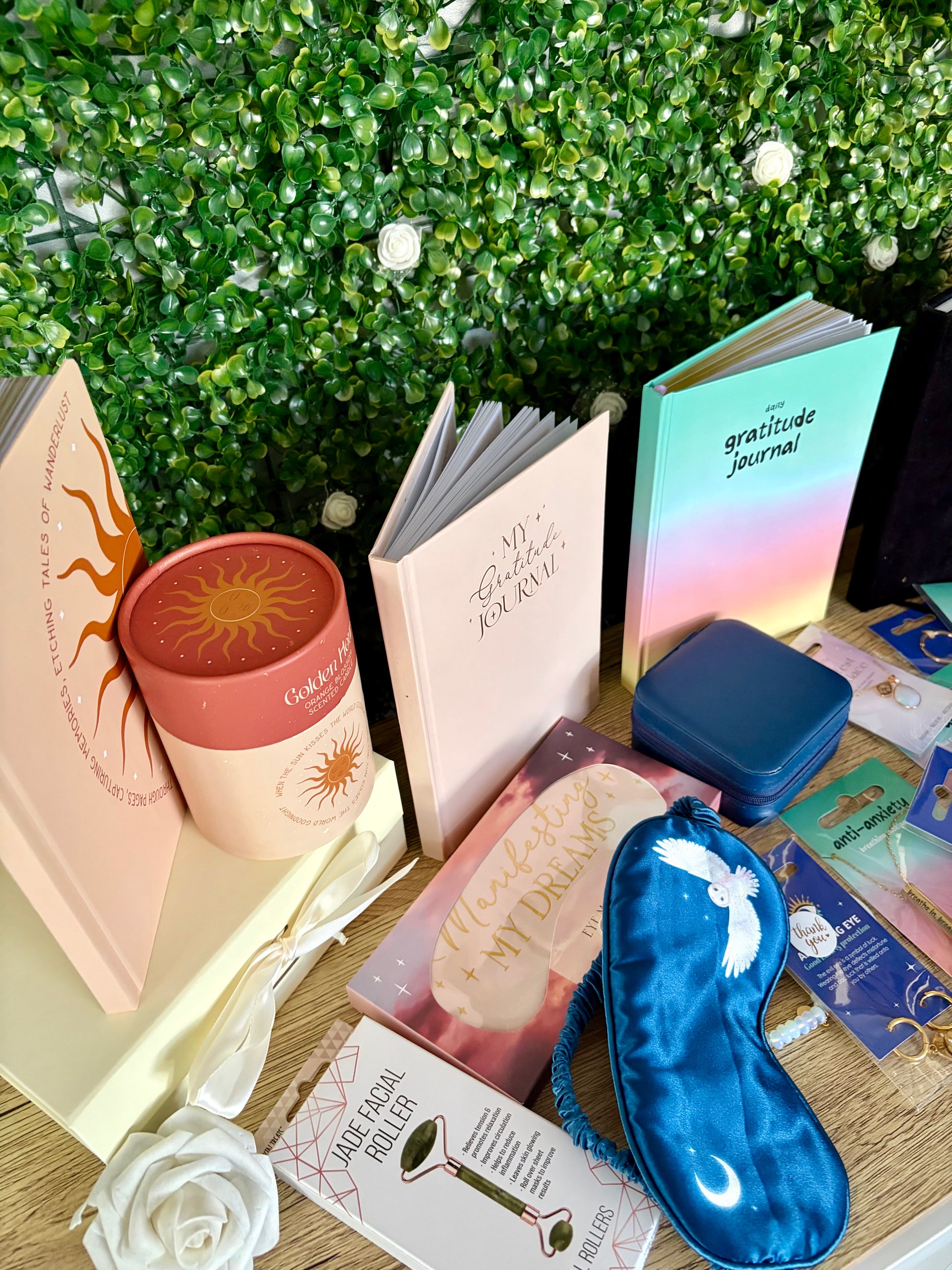 A styled flat-lay of various self-care and journaling products, including a blue satin sleep mask, a pink gratitude journal, a small blue jewelry box, and a pink sun-themed candle tin, arranged on a wooden surface in front of a green foliage background.