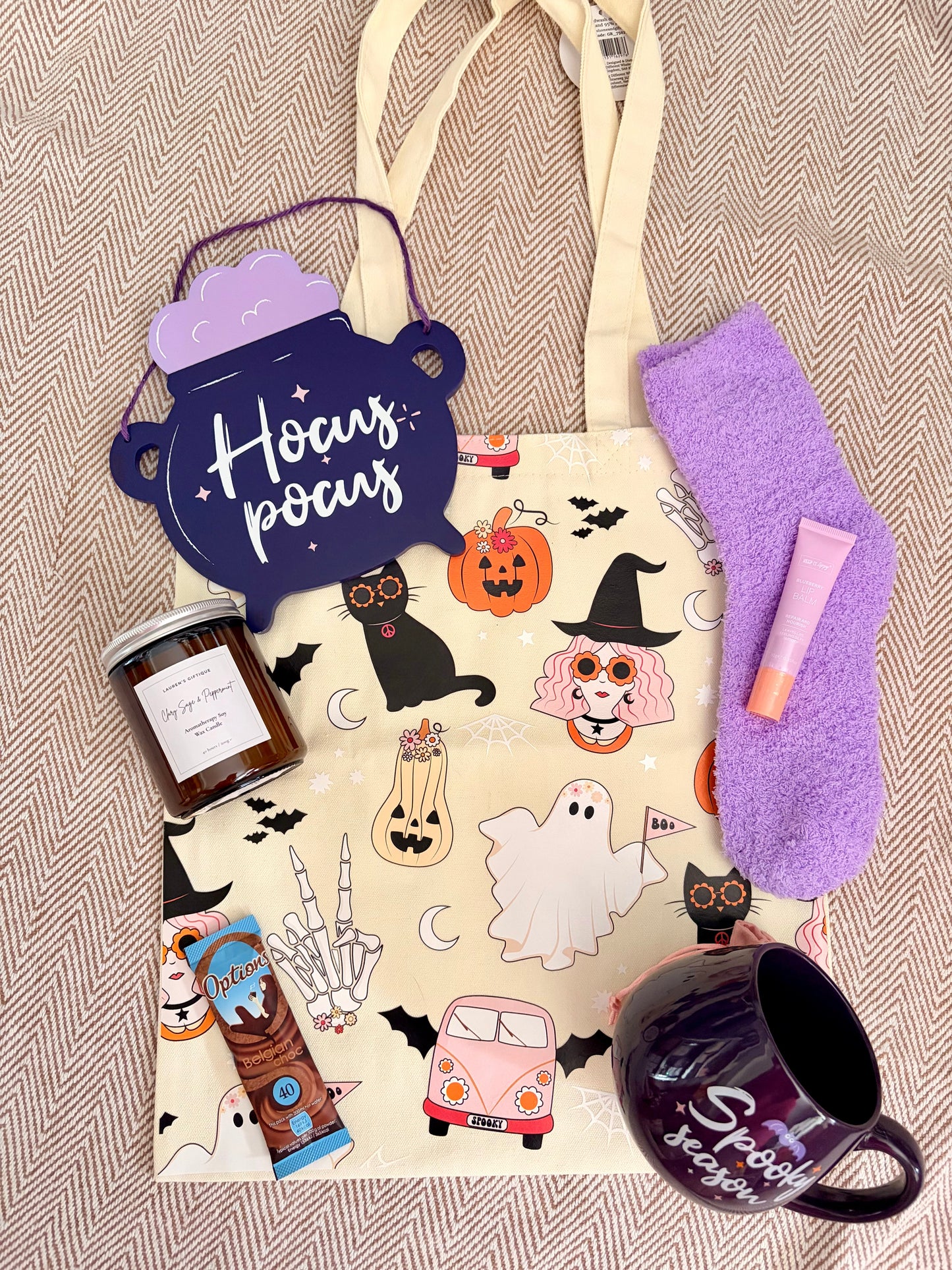 A cozy Halloween-themed gift set arranged on a beige herringbone fabric background. The set includes a cream tote bag with a 'Hocus Pocus' witch illustration print, a purple cauldron-shaped sign, a purple fuzzy sock, a pink lip balm tube, a candle jar, a purple mug, and a hot chocolate sachet.