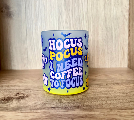 A frosted glass tumbler with a colorful graphic design featuring the text 'HOCUS POCUS I NEED COFFEE TO FOCUS' in blue and yellow lettering, set on a wooden surface against a wooden background.