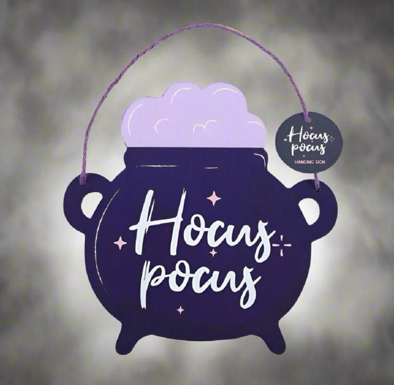 A hanging decorative cauldron sign with the words “Hocus Pocus” in white script, filled with light purple foam, suspended by a purple string against a soft gray blurred background. A small round tag on the right reads “Hocus Pocus Hanging Sign.