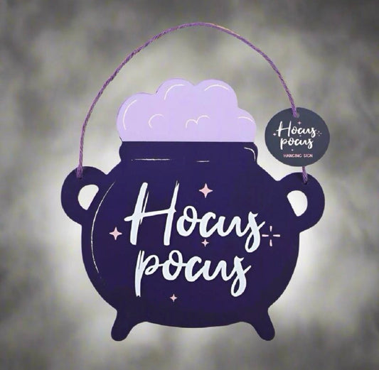A hanging decorative cauldron sign with the words “Hocus Pocus” in white script, filled with light purple foam, suspended by a purple string against a soft gray blurred background. A small round tag on the right reads “Hocus Pocus Hanging Sign.