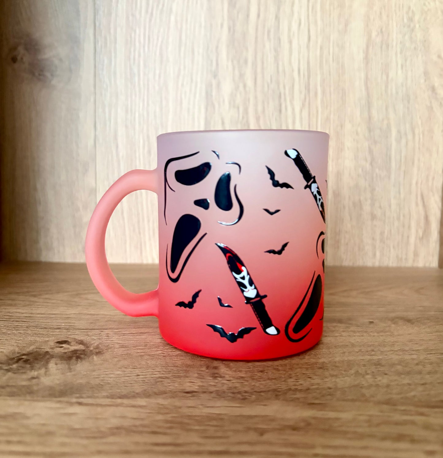 A frosted glass mug with a white-to-red gradient, decorated with black and white skull-and-crossbones and bat motifs, set on a wooden surface against a wood-paneled background.