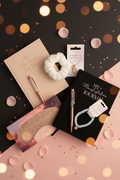Collection of self-care items including a pink notebook, journals, a necklace, and a bracelet on a textured surface.