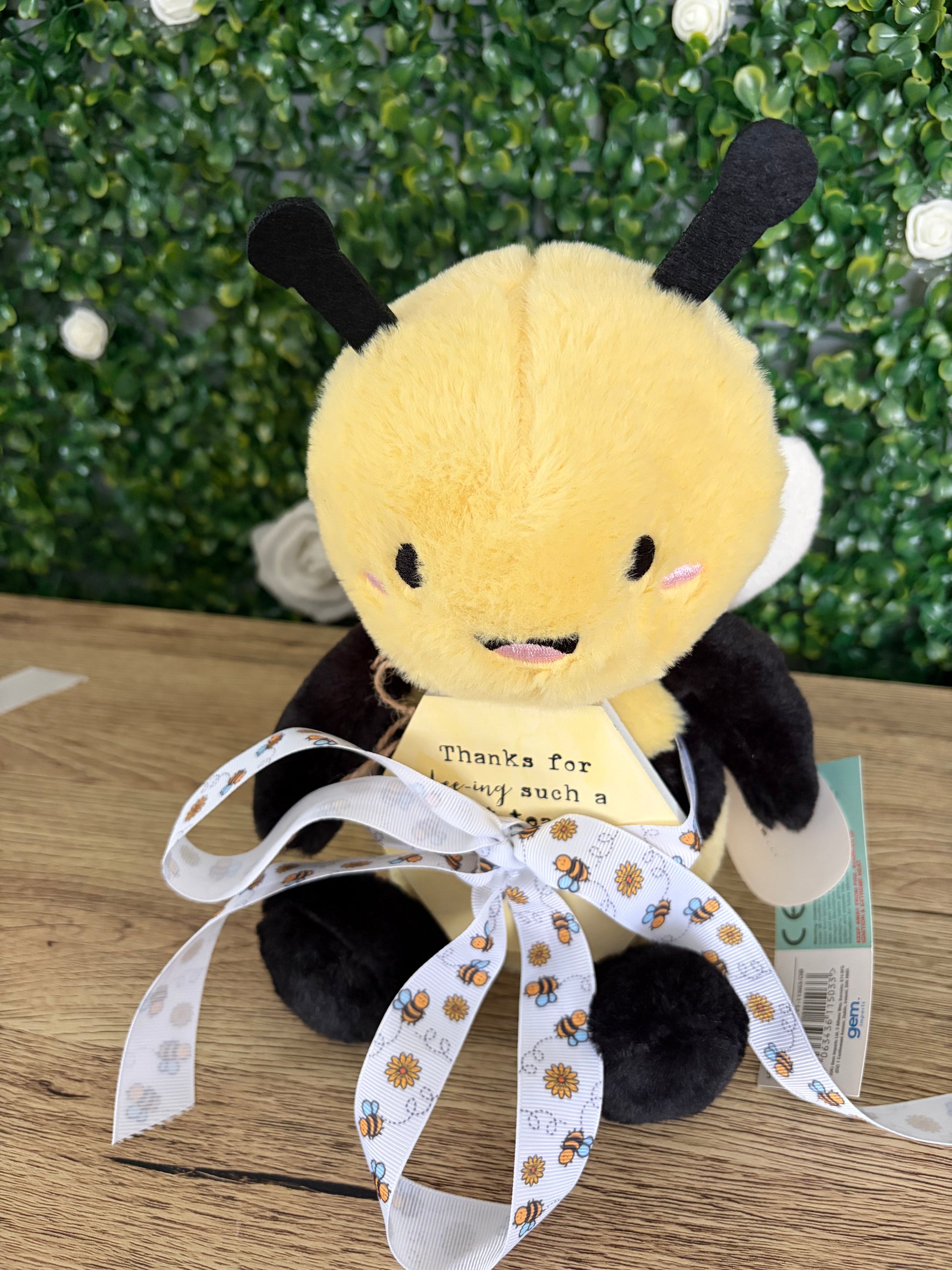 A yellow and black plush bee toy sitting on a wooden surface in front of a green leaf wall. The toy has black antennae, small white wings, and is tied with a white ribbon printed with bumblebees and sunflowers.