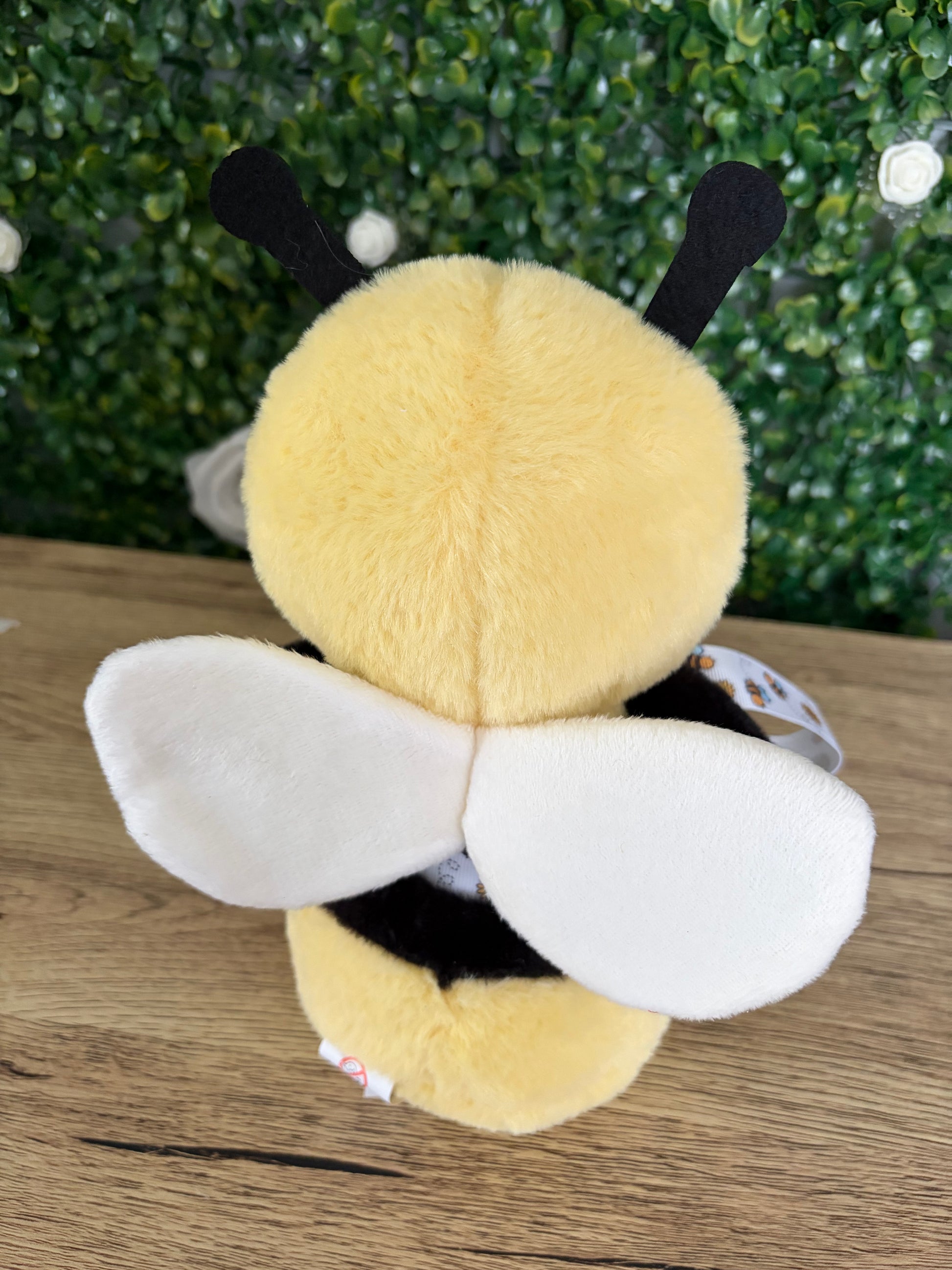 A yellow and black plush bee toy with white wings, photographed from the back on a wooden surface with a green hedge background.