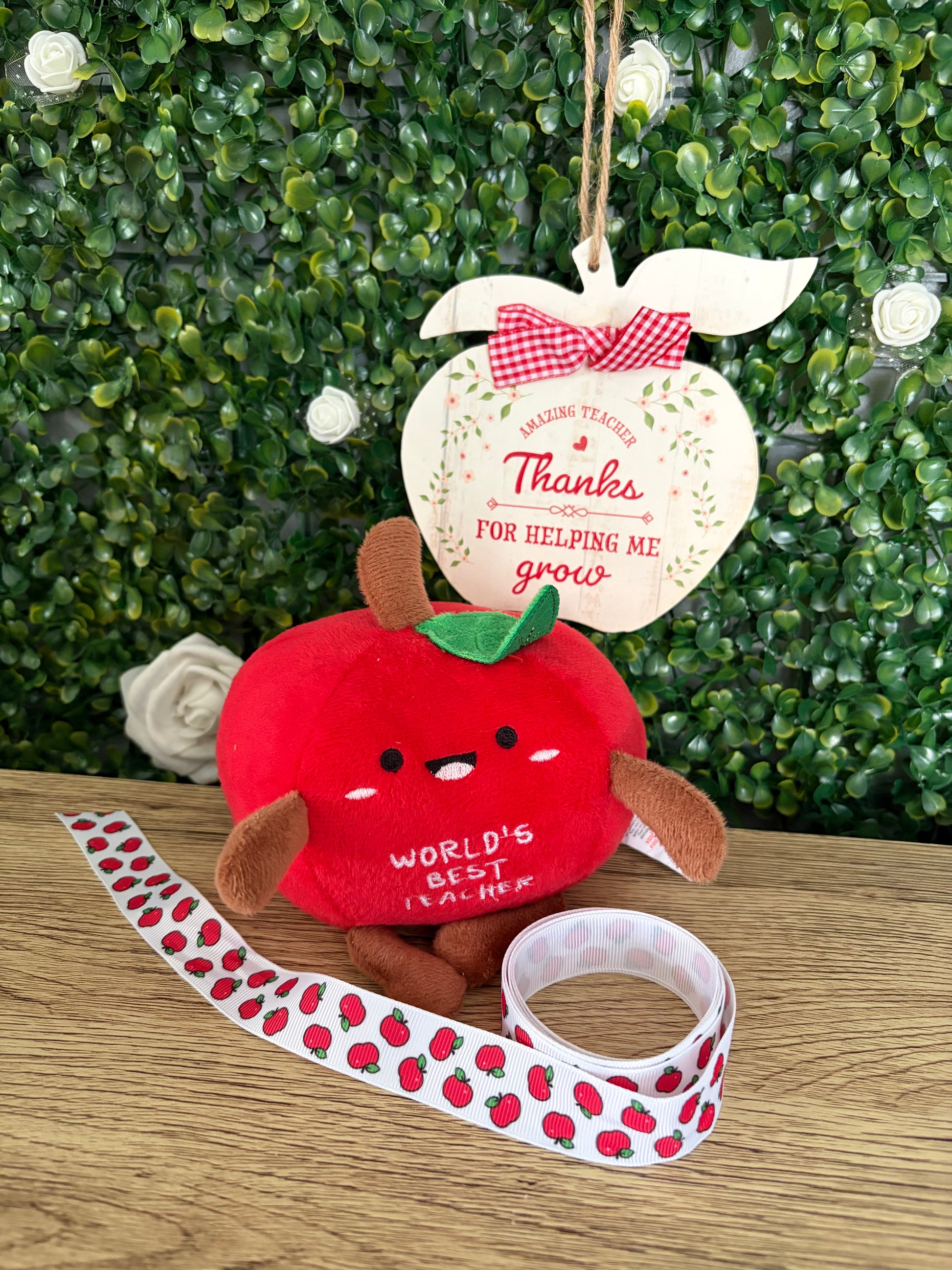 A red plush apple toy with brown arms and legs, a green stem, and embroidered text reading 'WORLD'S BEST TEACHER,' displayed on a wooden surface with a apple-patterned ribbon and a hanging wooden apple sign that says 'Thanks for helping me grow' in front of a green leafy backdrop.