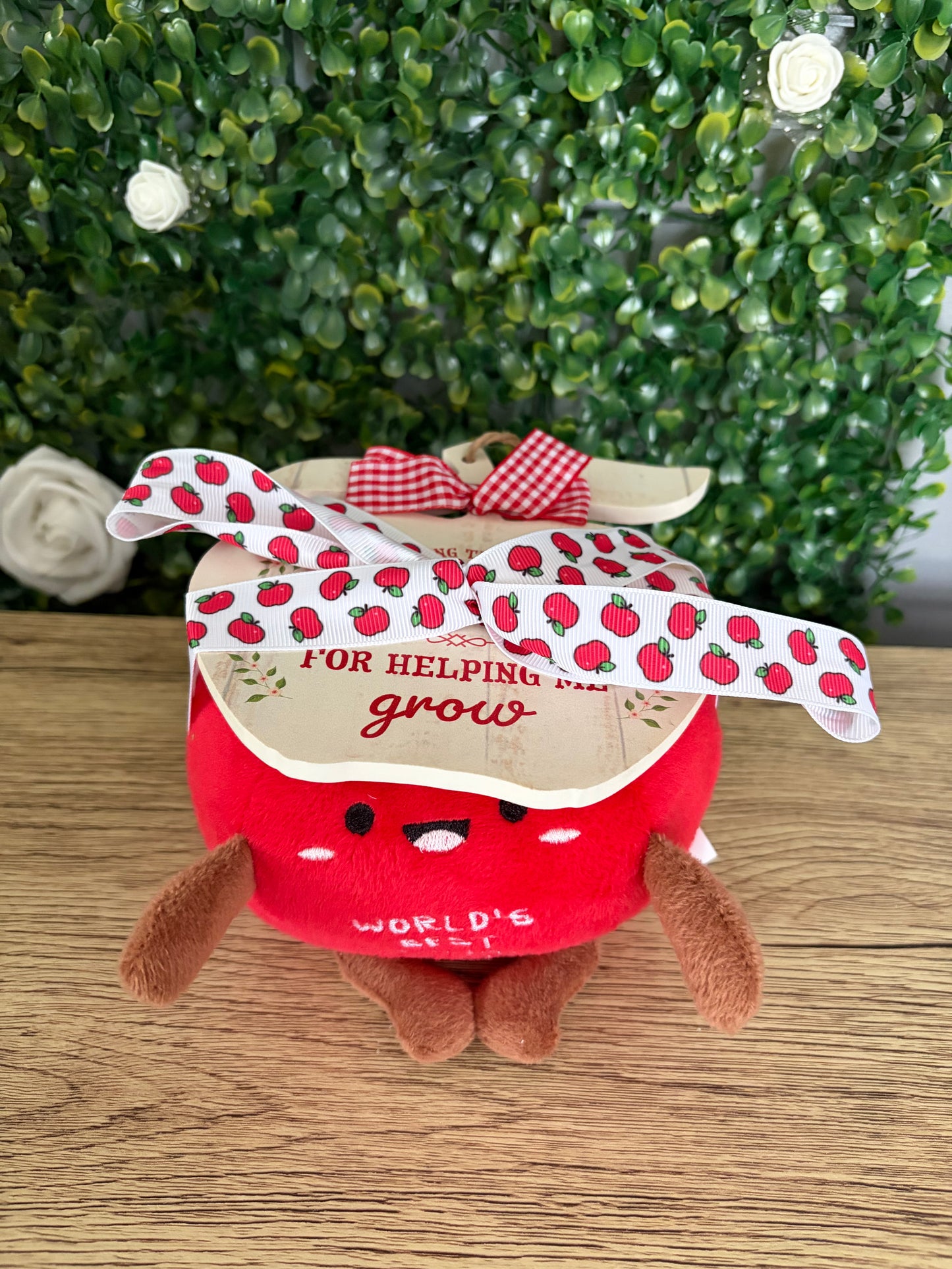 apple plush toy with brown arms and legs, with wooden apple sign tied on top with apple-printed ribbon on a wooden surface with green leafy background.
