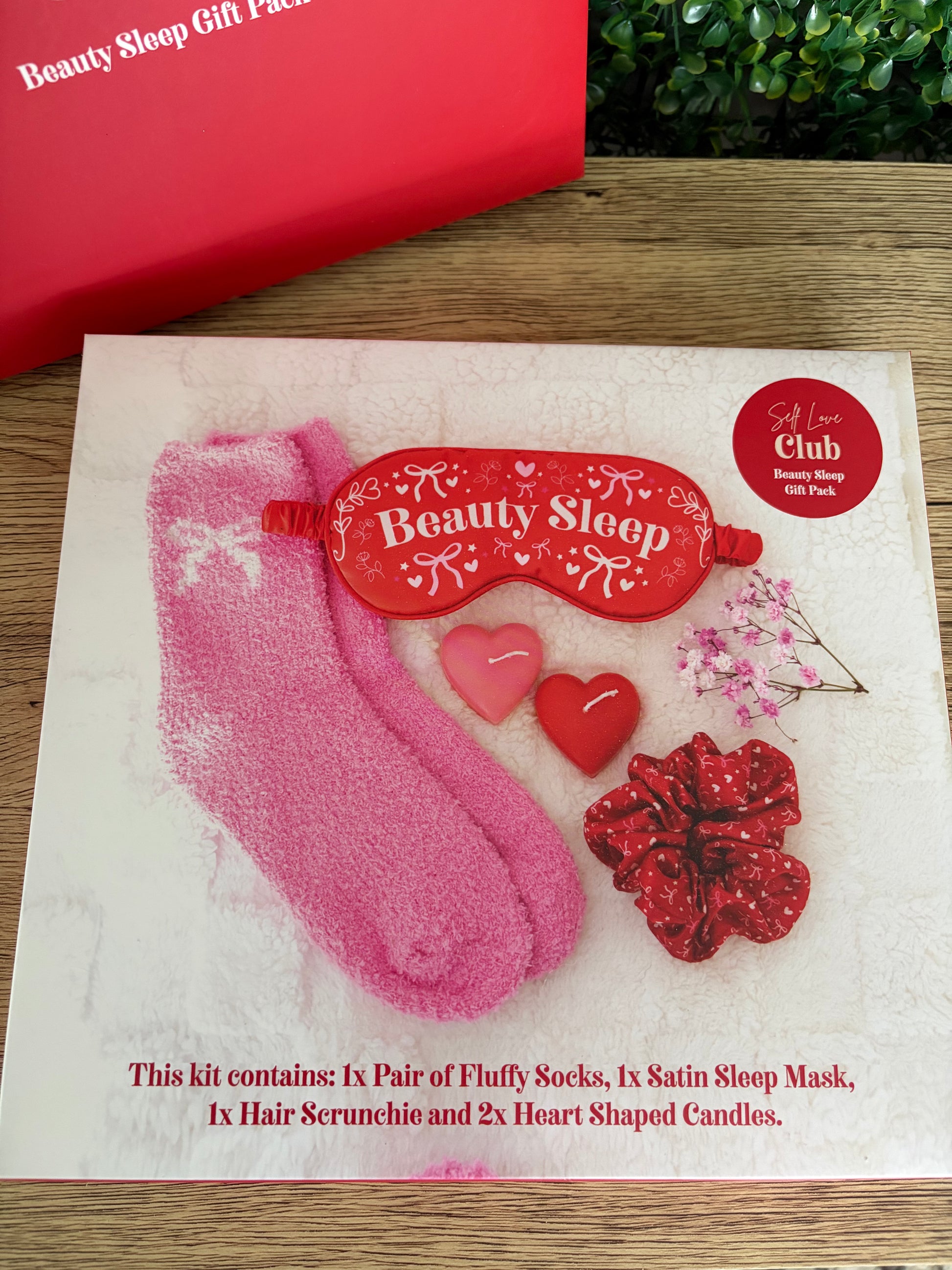 Beauty Sleep gift set on a white card, including pink fluffy socks, a red satin sleep mask, a red scrunchie, and two heart-shaped candles, displayed on a wooden surface with a red gift box partially visible.