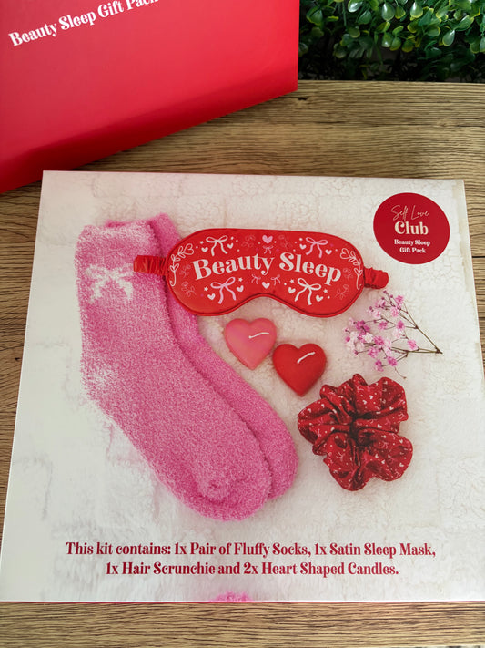 Beauty Sleep gift set on a white card, including pink fluffy socks, a red satin sleep mask, a red scrunchie, and two heart-shaped candles, displayed on a wooden surface with a red gift box partially visible.
