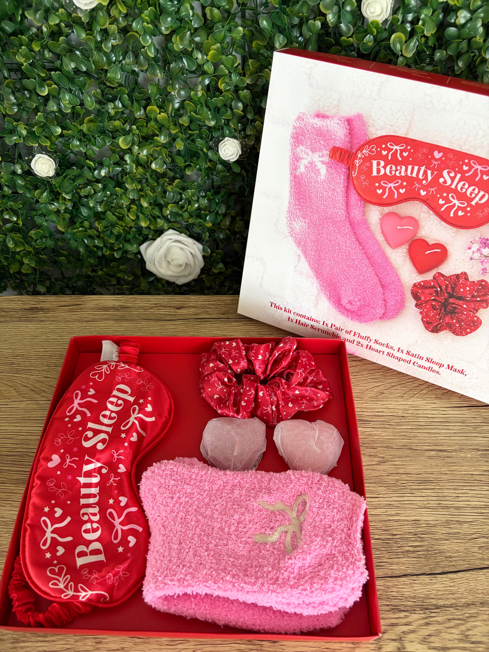 A pink and red beauty sleep gift set displayed in an open red box, featuring a red sleep mask with white script, pink socks, a satin red scrunchie, and two  heart-shaped candles. A printed box lid showing a pink sock and the words 'Beauty Sleep' sits behind the box on a wood surface with a green leafy backdrop.