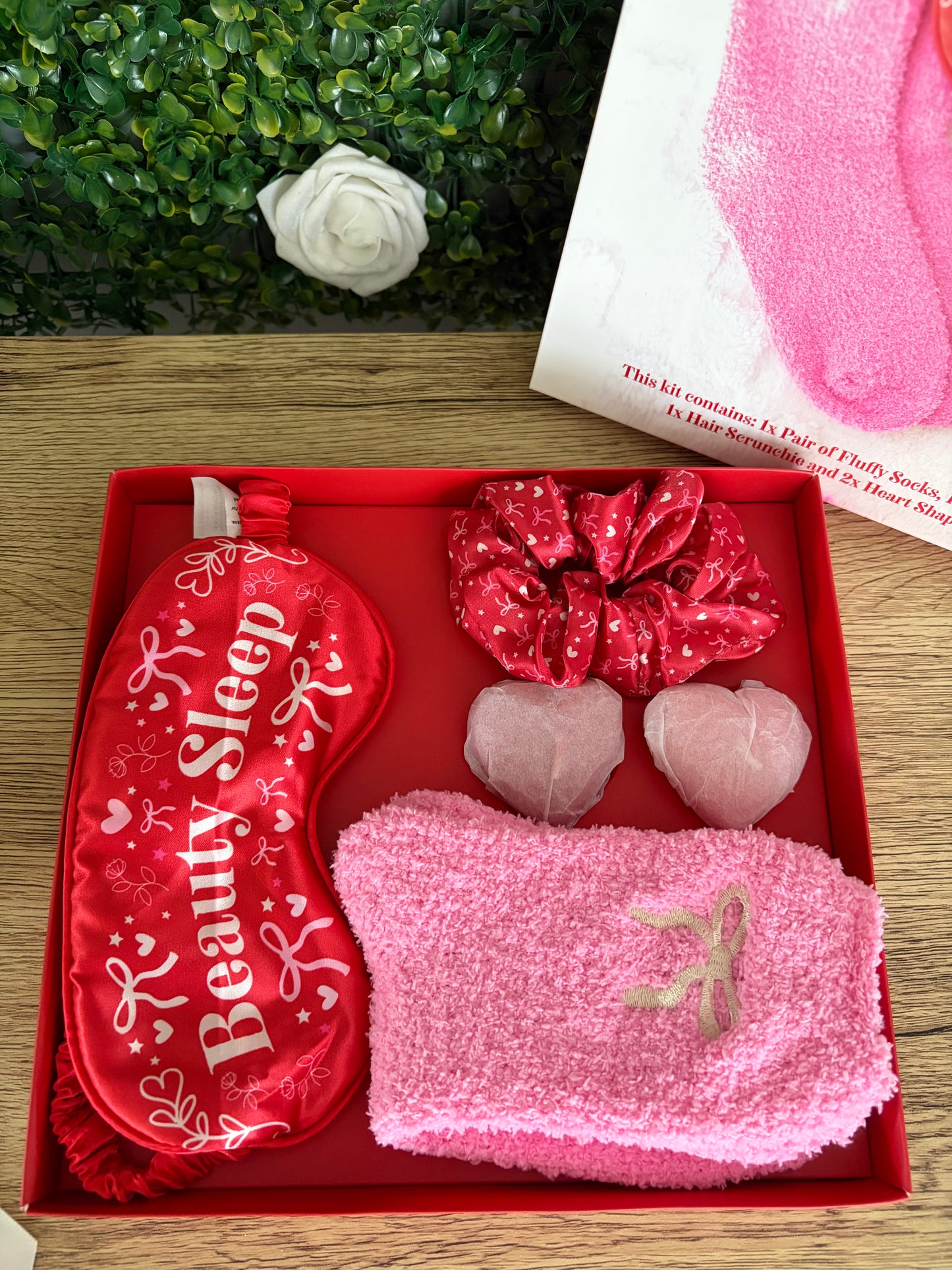 A beauty gift set in a red tray, featuring a red satin sleep mask with 'Beauty Sleep' printed in white, a red scrunchie, two  heart-shaped candles in tissue paper, and pink socks. A corner of lid and greenery are visible in the background on a wooden surface.