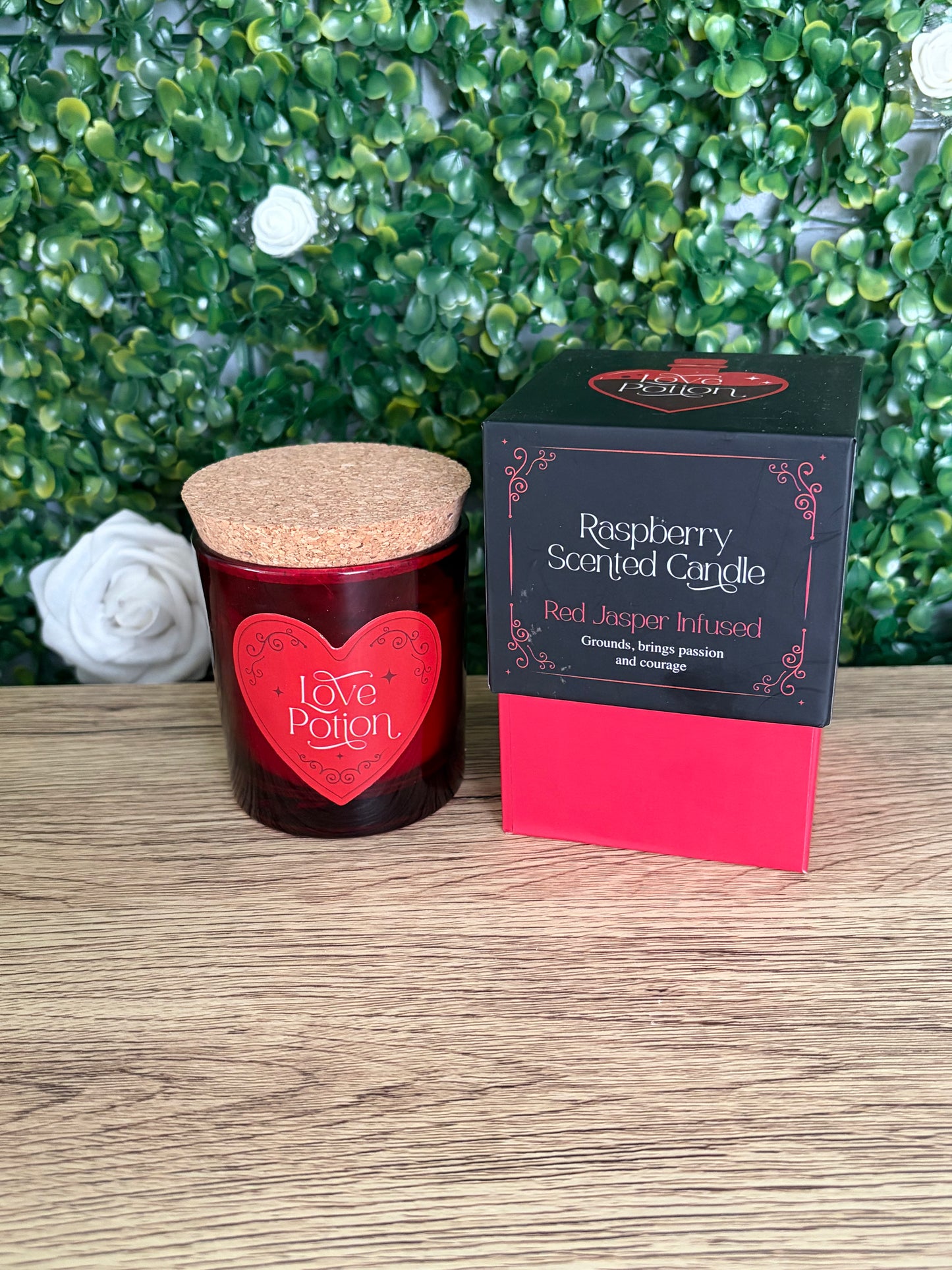 Raspberry scented candle box ed jasper infused with red glass jar and cork lid on a wooden surface with a green leafy backdrop