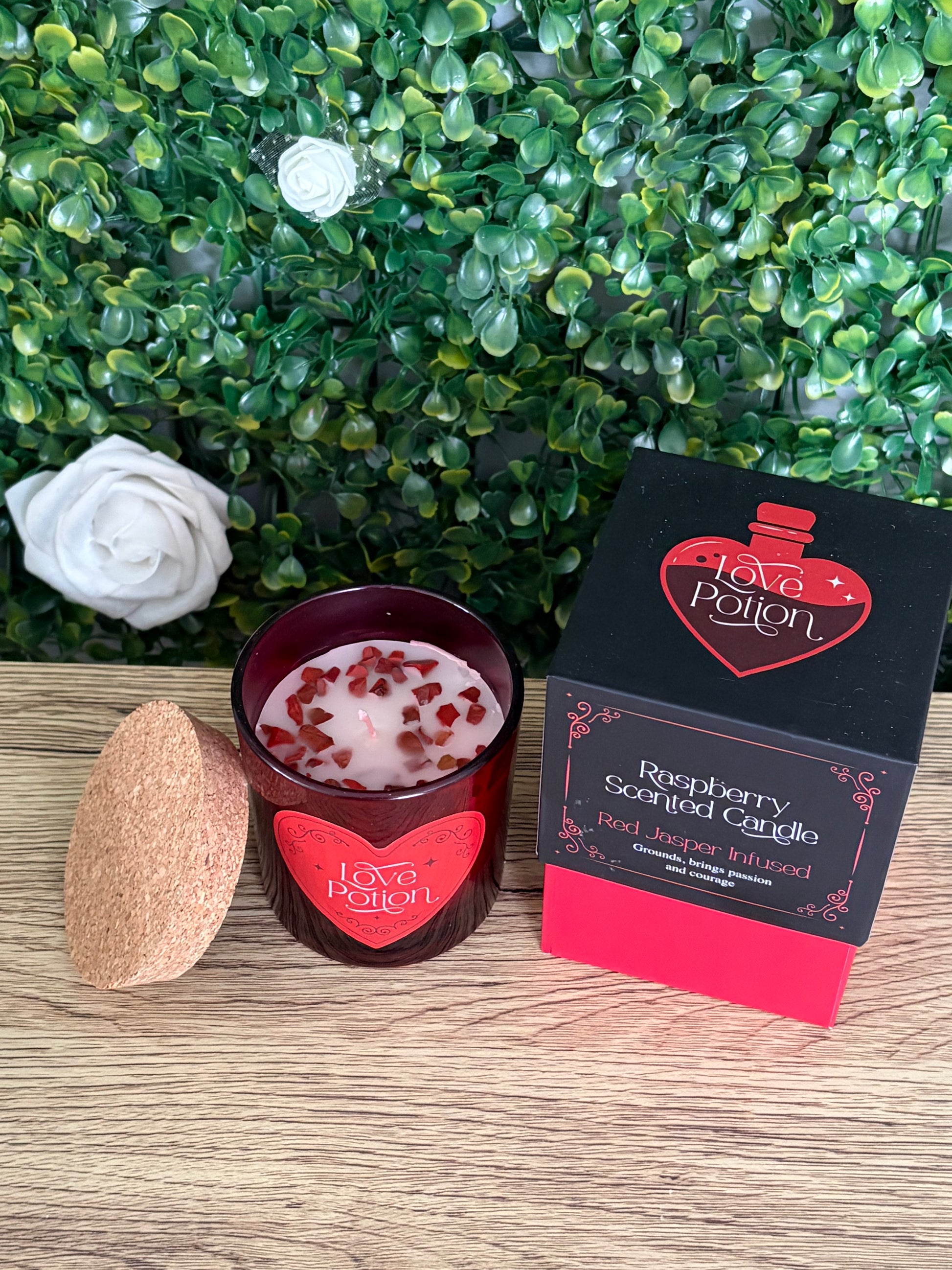 A Love Potion Raspberry Scented Candle shown with its black and red packaging box, alongside a cork lid, set against a green leafy background on a wooden surface.