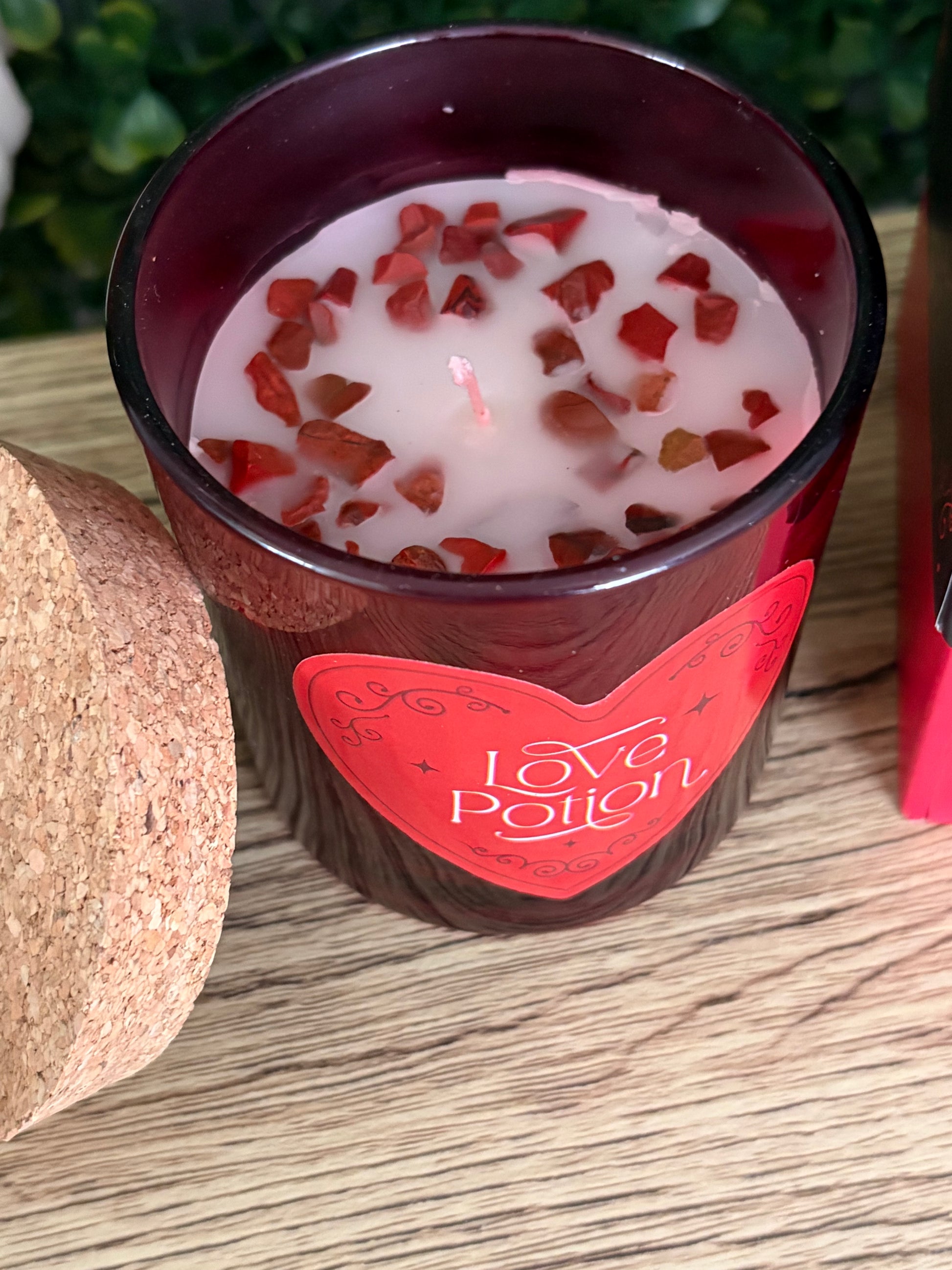 A dark red glass candle jar with a red heart-shaped label reading 'Love Potion' on a wooden surface, filled with white wax and topped with small red stone chips.