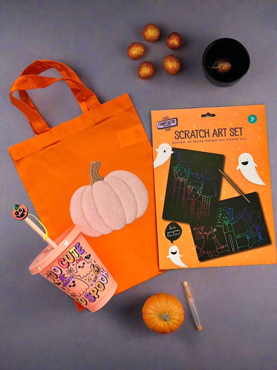 A Halloween-themed flat lay featuring a bright orange fabric tote bag with a white pumpkin design, a orange 'Too Cute, to Spook' cup, a packaged scratch art set, small pumpkins, and other seasonal props on a gray surface.