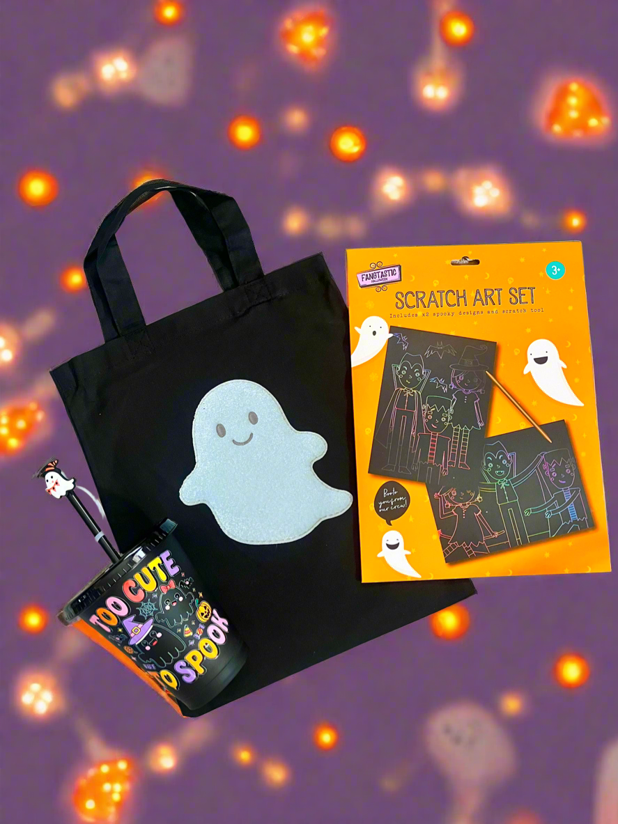 Halloween-themed gift set featuring a black canvas tote bag with a white ghost patch, a black cup with a ghost straw, and a Halloween Scratch Art Set in its orange packaging, arranged on a purple background with glowing orange bokeh lights.